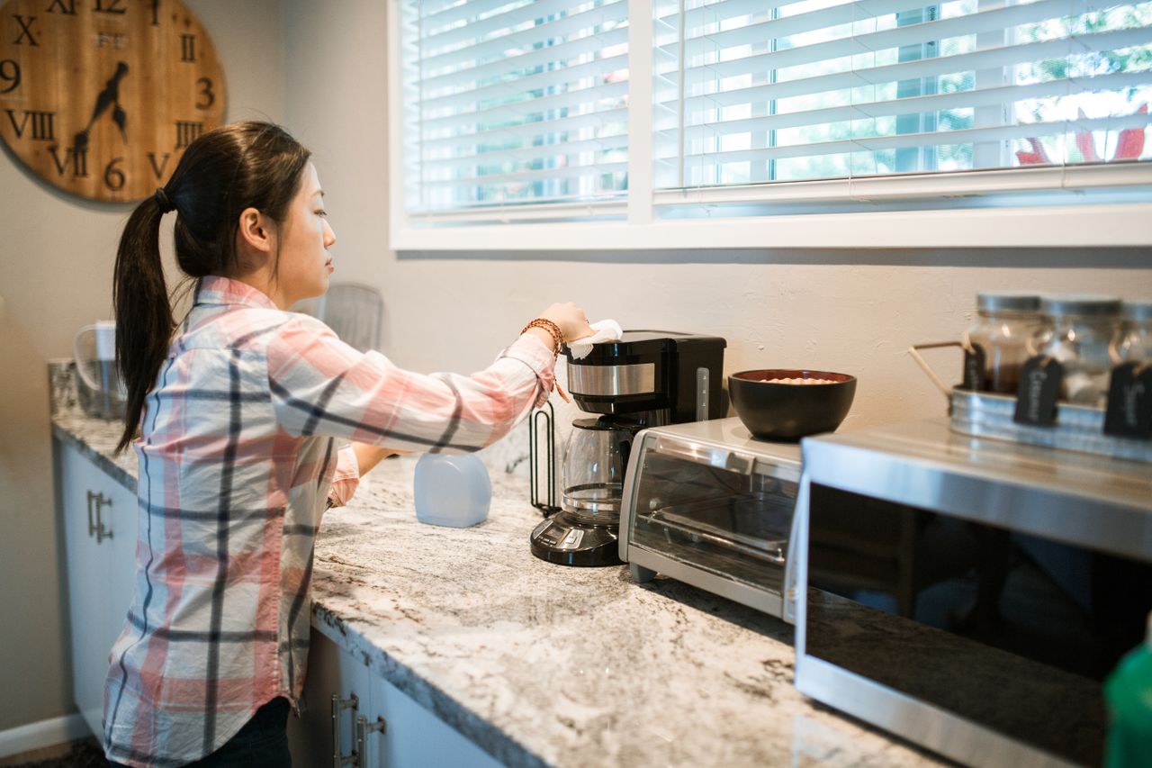 about-us Woman cleaning a coffee maker in a bright, modern kitchen with appliances.